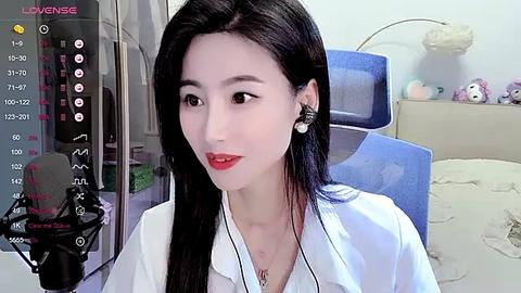 Media: Video of a young Asian woman with long black hair, wearing a white shirt, red lipstick, and earphones, sitting in a modern office. Background shows a blue chair, a white lamp, and a toy bunny.