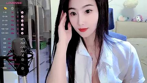 Media: A video of a fair-skinned Asian woman with long black hair, wearing a white shirt, sitting in front of a microphone, with a bedroom background featuring plush toys.