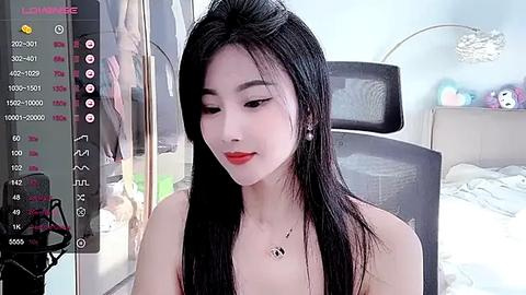 Media: Video of an East Asian woman with long black hair, fair skin, and red lipstick, wearing a black top, sitting in a modern bedroom with a grey chair and a bed in the background.