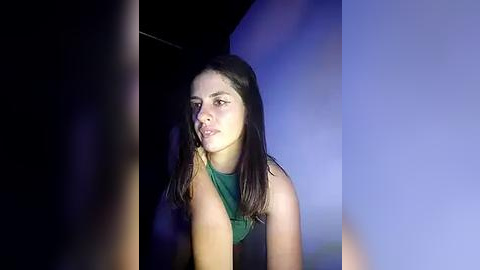 Media: A video of a young woman with long, straight, dark brown hair, wearing a green sleeveless top, looking serious, set against a dark, shadowy background with a blue light accent.