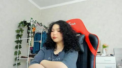 Media: A video of a young woman with curly black hair, wearing a grey T-shirt, sitting in a red and black gaming chair. The room has a white wall, a green leafy plant, and a shelving unit with clothes.