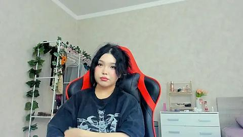 Media: Video of an Asian woman with long black hair, wearing a black graphic t-shirt, sitting in a red gaming chair in a simple, light-colored room with a white shelving unit and potted plants.