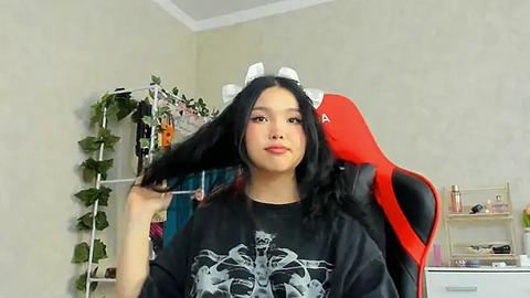 Media: Video of an Asian woman with long black hair, wearing a black T-shirt with a skull graphic, and a cow headband, seated in a red gaming chair in a simple, beige-walled room with a green plant and bookshelf.