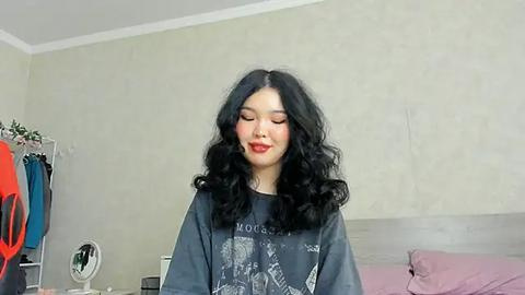 Media: Video of a young Asian woman with long, voluminous black hair, wearing a gray graphic tee, red lipstick, standing in a modern bedroom with muted beige walls, white trim, and a purple bed.