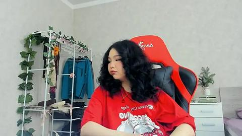 Media: Video of a young woman with wavy black hair, wearing a red T-shirt with a white graphic, seated on a red gaming chair, surrounded by a cluttered room with a white dresser and green plants.