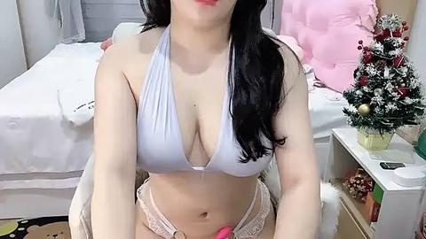 Media: Video of an Asian woman with long black hair, wearing a white halter top and pink lace panties, sitting on a white chair in a bedroom with a white bed, pink pillows, and a Christmas tree.