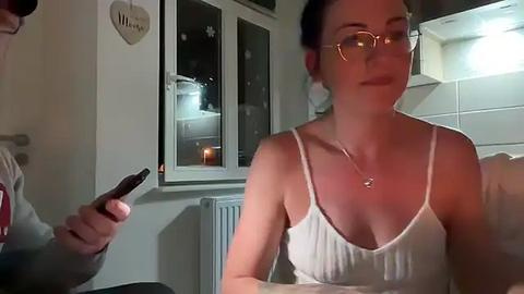 Media: A video of a fair-skinned woman with glasses and white spaghetti straps, holding a phone in a dimly-lit room with a window, radiator, and a heart-shaped wall plaque.