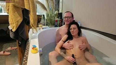 Media: Video of a nude couple in a bathtub, a man with glasses and a tattooed arm, holding a sex toy, and a woman with medium-sized breasts, in a modern bathroom.