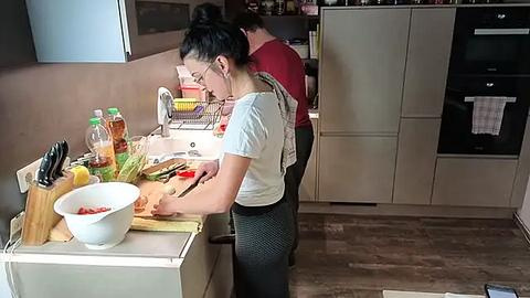 Media: Video of two women cooking in a modern kitchen; one with a bun, chopping vegetables on a wooden counter, the other in a red top, reaching for a pot.