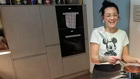 Media: A video shows a smiling woman with glasses and tattoos, wearing a Mickey Mouse T-shirt and apron, cooking in a modern kitchen with beige cabinets and a microwave.