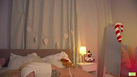 Media: Video of a cozy, dimly lit bedroom with a white bed, heart garland, red-striped stocking, and a nightstand lamp.