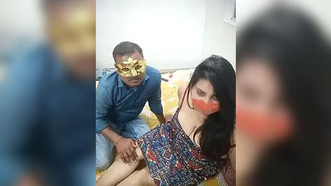 Media: A video of a young woman with long black hair and a red mask, wearing a colorful patterned top, being held down by a man in a blue shirt and a golden mask on a wooden floor in a room.