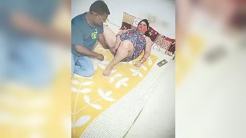 Media: A video shows a chubby, pale-skinned woman in a floral bikini lying on a yellow beach towel with white birds, while a black man in glasses and a T-shirt massages her thigh.