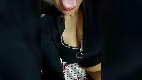 Media: Video of a woman with fair skin, wearing a black shirt with a zipper, licking her lips, with a patterned white and black fabric visible in the background.