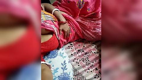 Media: Video of a woman lying on a bed in a pink sari, with a red blouse visible, surrounded by colorful bedcovers. The woman's hand is placed on her abdomen.