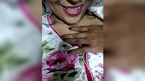 Media: Video of a dark-skinned woman with a wide smile, wearing a pink floral blouse and a gold chain, touching her chest with manicured nails, blurred background.