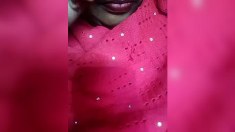 Media: Video of a woman's face partially covered by a vibrant pink fabric with a textured, dotted pattern. The fabric appears to be draped over her shoulders, highlighting her dark complexion and full lips.