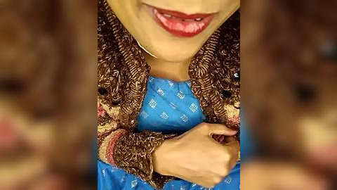 Media: A close-up video of a woman with fair skin, wearing a blue dress adorned with gold embroidery and a brown shawl, smiling with red lipstick, her hands clasped in front of her.