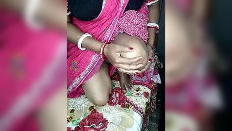 Media: Video of an Indian woman in a bright pink saree with intricate white patterns, kneeling on a floral-patterned mat, gently cradling her baby in her arms.