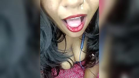 Media: Video of a young woman with light skin and long, dark hair, wearing a red patterned top and a nose piercing, her mouth open wide, tongue out, and a blue headphone cord around her neck.