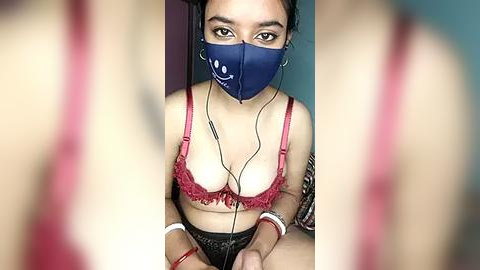 Media: Video of a young woman with medium skin tone, wearing a blue mask and red lace bra, sitting on a bed, hands holding headphones, blurred background.