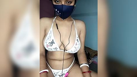 Media: Video of a young woman with medium skin tone, wearing a white bikini with colorful patterns, a blue face mask, and headphones. She sits on a bed with muted teal and pink sheets, in a dimly lit room.