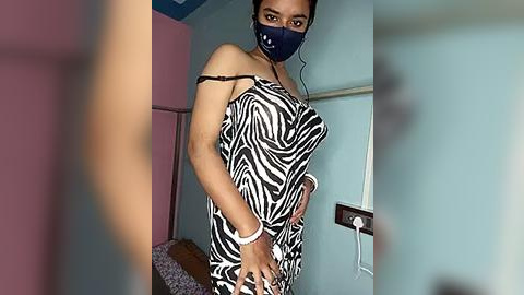 Media: Video of a woman in a zebra-print dress with a black mask, standing in a dimly lit room with pastel walls and a bed.