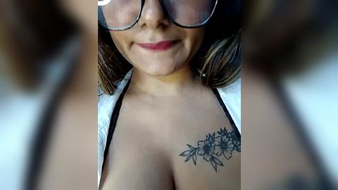 Media: Video of a smiling, light-skinned woman with brown hair, wearing black-rimmed glasses and a low-cut top, showcasing a black floral tattoo on her collarbone.