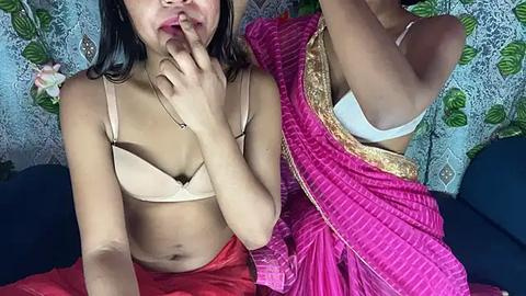 Media: Video of two women, one in a beige bra, the other in a pink sari, with a white bra visible, against a floral-patterned curtain background.