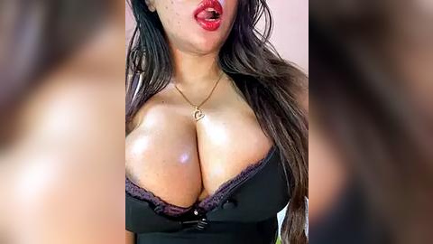 Media: Video of a woman with long, dark hair and medium skin tone, wearing a black lace bra that accentuates her large, round breasts. She has red lipstick and a gold necklace with a heart pendant.