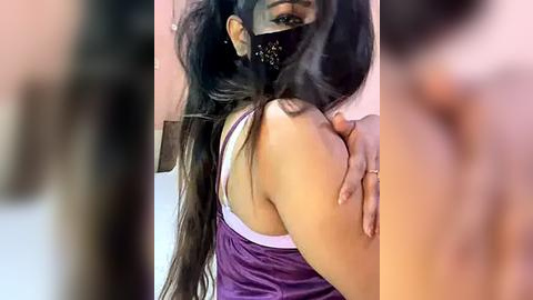 Media: A video of a woman with long black hair, wearing a purple tank top and black mask, with a blurred background, showing her side profile.