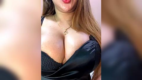 Media: Video of a light-skinned woman with long brown hair, wearing a black, low-cut, lacy bra, and a gold necklace with a heart pendant, showing cleavage prominently. Background is blurred and indistinct.