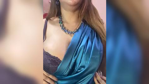 Media: Video of a light-skinned woman with long brown hair, wearing a teal silk sari with a lace black bra visible, a turquoise necklace, and a blurred background.