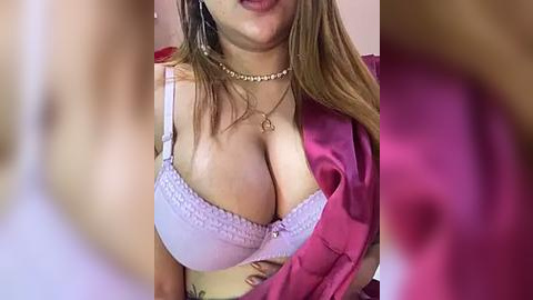 Media: Video of a woman with long brown hair, wearing a white lace bra, a pink satin robe, and a pearl necklace. Her cleavage is prominent, and she has a tattoo on her side.