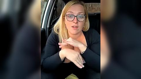 Media: Video of a blonde woman with glasses, medium build, wearing a black top, hands clasped in a car, blurred figures in the foreground.