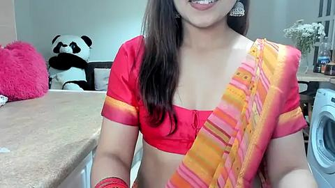 Media: Video of a smiling Indian woman in a vibrant red and orange striped saree, wearing large silver earrings, standing in a modern kitchen with a panda toy and a washing machine.