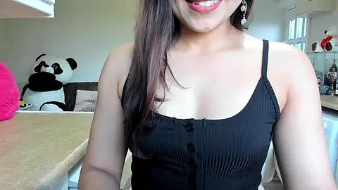 Media: Video of a smiling, light-skinned Asian woman with long, straight dark hair, wearing a black, ribbed, spaghetti-strapped top. Background features a panda plush toy, kitchen counter, and kitchen decor.