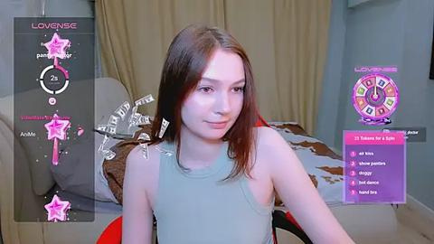 Media: Video of a fair-skinned, long-haired young woman with a small bust, sitting on a red chair in a bedroom, wearing a light green tank top.