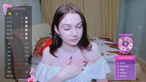 Media: A video of a young Caucasian woman with fair skin and dark hair, wearing a white polka-dot off-shoulder top, sitting indoors.