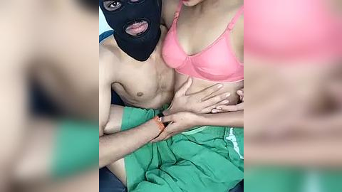 Media: A video shows a shirtless man with a black mask, green shirt, and a woman in a pink bra, both with light skin. They are seated intimately, hands intertwined.