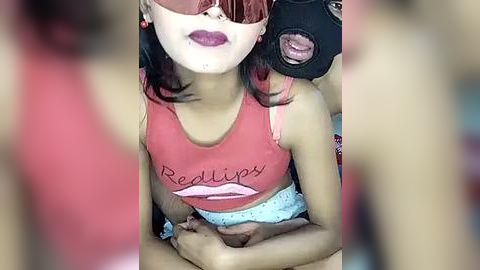 Media: A video of a woman with a red and pink hairdo, wearing a red tank top with \"Red Lips\" printed on it, and a black mask with pink lipstick marks, holding a black camera.