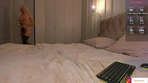 Media: A video of a dimly lit bedroom showing a nude blonde woman standing in a doorway, a smartphone on the bed with a virtual reality app open, and a plush headboard.