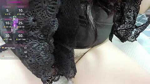 Media: A close-up video of a woman with fair skin and dark hair, wearing black lace lingerie and a black lace robe, lying on a bed with white sheets. The image includes a digital time display with a pink background.