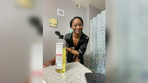 Media: Video of a smiling Black woman with braids in a bathroom, holding a bottle of olive oil. Gray walls, blue shower curtain, and a speckled countertop.