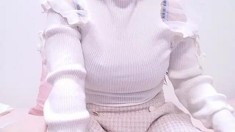 Media: Video of a woman wearing a white, ribbed, long-sleeve top with frilled shoulders and a high-waisted, pleated skirt. Her skin tone is light, and she is seated in a room with a white background and a pink object to the left.