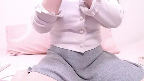 Media: Video of a fair-skinned person with light hair, wearing a white button-up shirt and gray shorts, sitting on a pink bed with a pillow and blanket, creating a soft, pastel aesthetic.