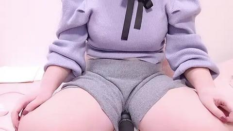 Media: Video of a fair-skinned woman in a light purple sweater and gray high-waisted shorts, sitting on a bed with white sheets. She is partially visible, with hands resting on her thighs.