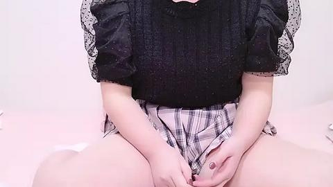 Media: Video of a fair-skinned woman seated on a bed, wearing a black lace top with puffed sleeves and a plaid skirt, her hands modestly covering her crotch.