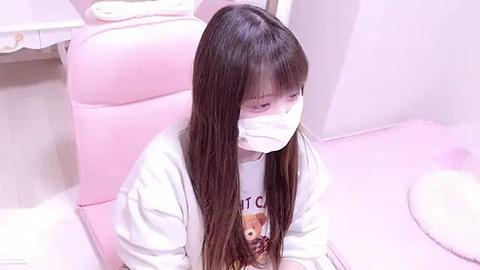 Media: Video of an Asian girl with long brown hair, wearing a white mask, white t-shirt, and pink chair in a pastel-toned room.