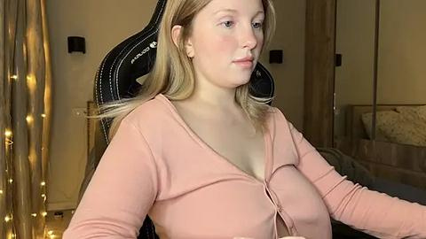 Media: Video of a Caucasian woman with blonde hair, wearing a pink cardigan, seated in a gaming chair, with a beige curtain and a bed in the background.